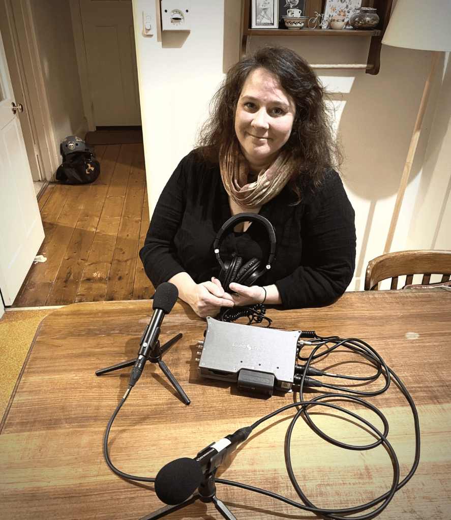A woman sitting at a table with recording equipment - two microphones and a sounds unit. She has dark long hair, is wearing a black top and is holding a set of headphones.